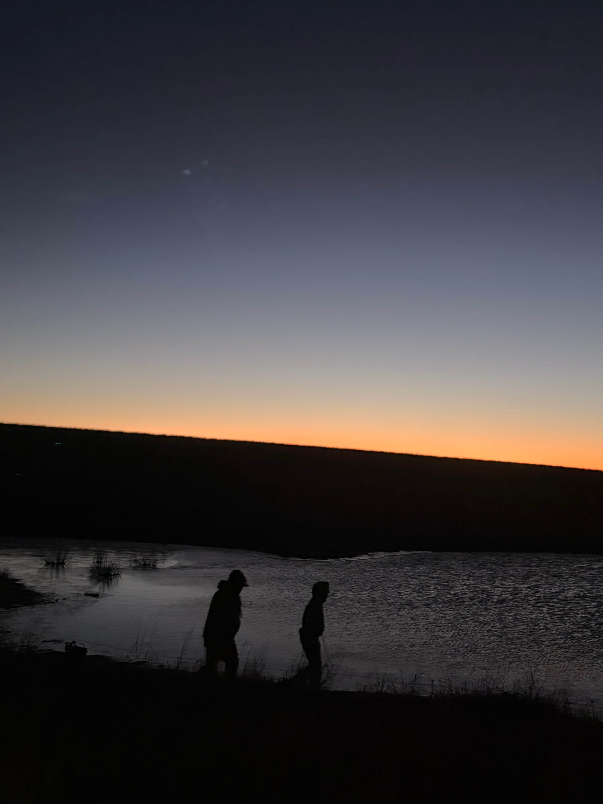 silhouettes of two hunters walking near water at dusk