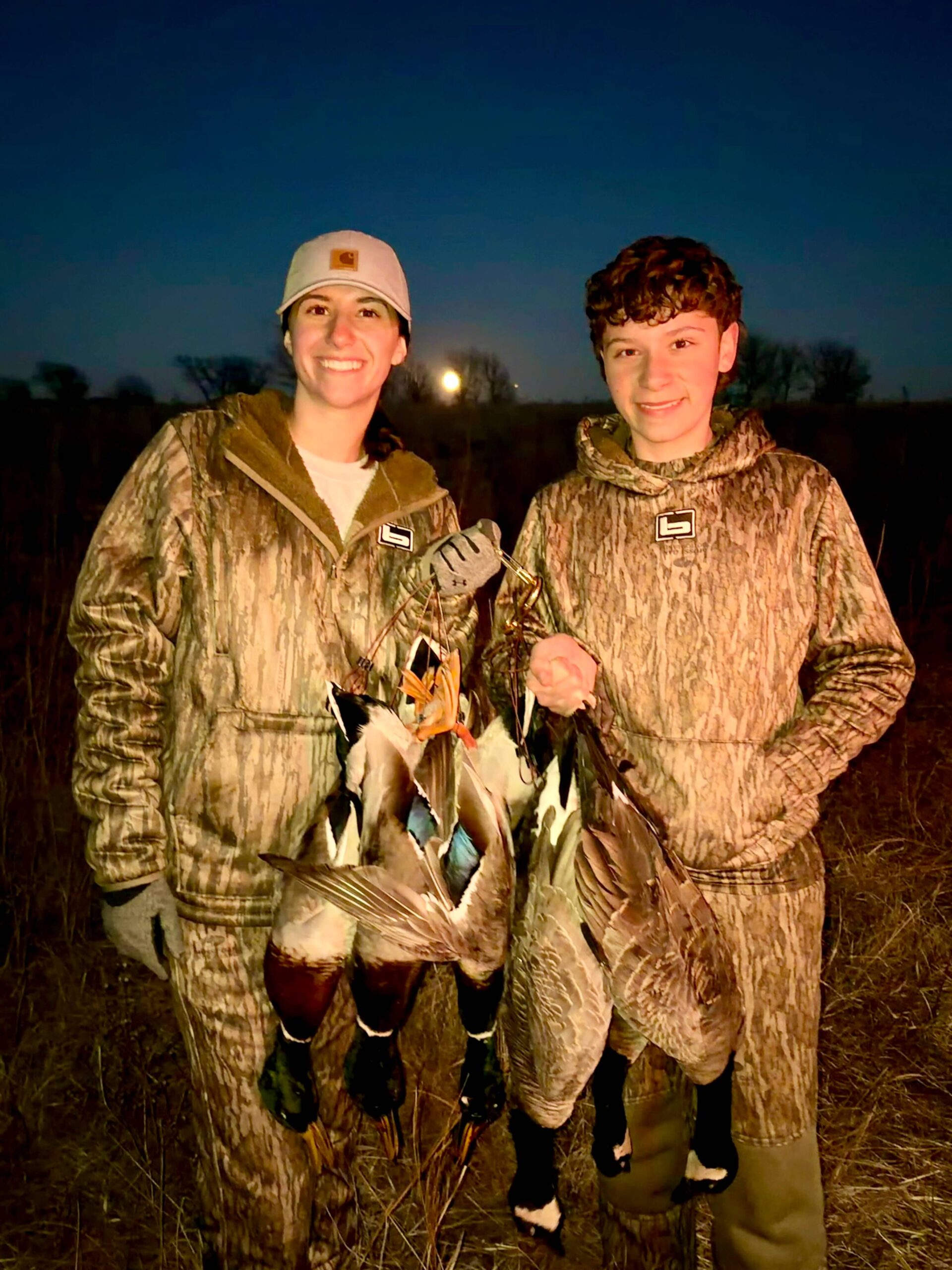 two hunters holding a strap of ducks at dusk